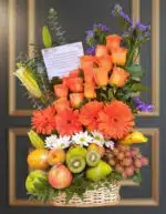 Arreglo floral con rosas y gerberas naranjas, margaritas blancas y flores moradas acompañado de canasta de frutas con manzanas, peras, kiwi, uvas, plátano y otras frutas frescas