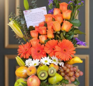 Arreglo floral con rosas y gerberas naranjas, margaritas blancas y flores moradas acompañado de canasta de frutas con manzanas, peras, kiwi, uvas, plátano y otras frutas frescas