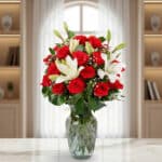 Arreglo floral con rosas rojas y lirios blancos en un jarrón de cristal sobre una mesa de mármol, con ventana y estanterías de fondo desenfocado