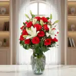 Arreglo floral con rosas rojas y lirios blancos en un jarrón de cristal sobre una mesa de mármol, con ventana y estanterías de fondo desenfocado