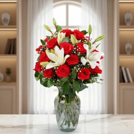 Arreglo floral con rosas rojas y lirios blancos en un jarrón de cristal sobre una mesa de mármol, con ventana y estanterías de fondo desenfocado