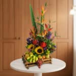 Arreglo floral tropical con girasoles, aves del paraíso, flores moradas y rojas combinado con frutas como uvas, manzanas y plátanos pequeños en base de bambú sobre mesa blanca
