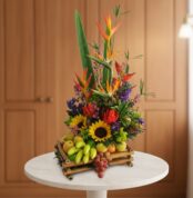 Arreglo floral tropical con girasoles, aves del paraíso, flores moradas y rojas combinado con frutas como uvas, manzanas y plátanos pequeños en base de bambú sobre mesa blanca