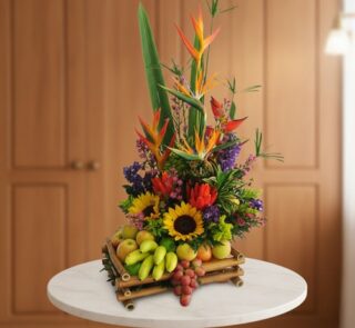 Arreglo floral tropical con girasoles, aves del paraíso, flores moradas y rojas combinado con frutas como uvas, manzanas y plátanos pequeños en base de bambú sobre mesa blanca