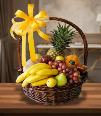 canasta de mimbre con plátanos, uvas, piña, peras y otras frutas variadas decorada con gran lazo amarillo sobre mesa de madera en interior elegante