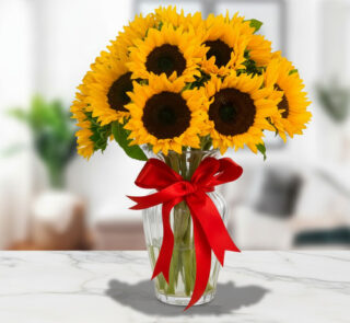 Arreglo de girasoles amarillos en un florero de cristal transparente decorado con un lazo rojo sobre una mesa de mármol