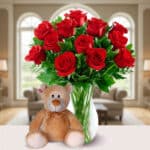 Jarrón de cristal con ramo de rosas rojas y hojas verdes junto a un oso de peluche marrón claro sobre una mesa blanca en una sala de estar luminosa