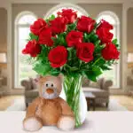 Jarrón de cristal con ramo de rosas rojas y hojas verdes junto a un oso de peluche marrón claro sobre una mesa blanca en una sala de estar luminosa