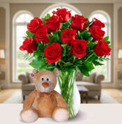 Jarrón de cristal con ramo de rosas rojas y hojas verdes junto a un oso de peluche marrón claro sobre una mesa blanca en una sala de estar luminosa