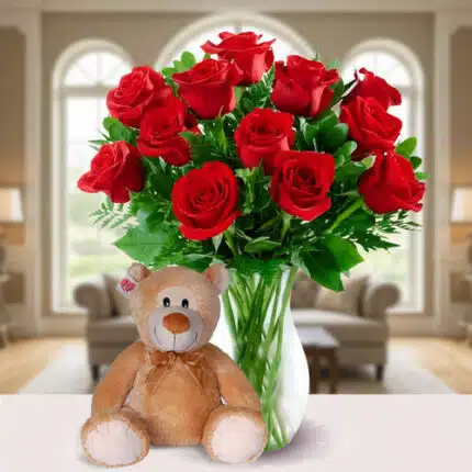 Jarrón de cristal con ramo de rosas rojas y hojas verdes junto a un oso de peluche marrón claro sobre una mesa blanca en una sala de estar luminosa