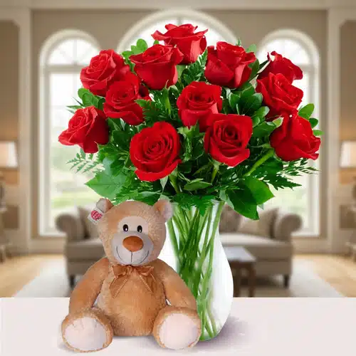 Ramo de rosas rojas con oso de peluche Jarrón de cristal con ramo de rosas rojas y hojas verdes junto a un oso de peluche marrón claro sobre una mesa blanca en una sala de estar luminosa