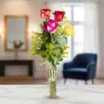 Ramo vertical de rosas rojas, rosas, blancas y amarillas con follaje verde en jarrón de cristal estrecho sobre mesa de mármol, con sala de estar desenfocada al fondo