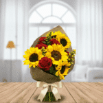 Ramo de flores con girasoles amarillos y rosas rojas envuelto en papel kraft y lazo crema, apoyado sobre mesa de madera con sala luminosa de fondo