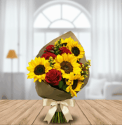 Ramo de flores con girasoles amarillos y rosas rojas envuelto en papel kraft y lazo crema, apoyado sobre mesa de madera con sala luminosa de fondo