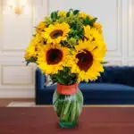 Arreglo de girasoles amarillos con follaje verde en un jarrón de cristal con cinta naranja, colocado sobre una mesa de madera frente a un sofá azul y una pared elegante