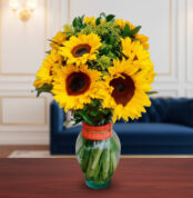 Arreglo de girasoles amarillos con follaje verde en un jarrón de cristal con cinta naranja, colocado sobre una mesa de madera frente a un sofá azul y una pared elegante