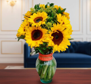 Arreglo de girasoles amarillos con follaje verde en un jarrón de cristal con cinta naranja, colocado sobre una mesa de madera frente a un sofá azul y una pared elegante