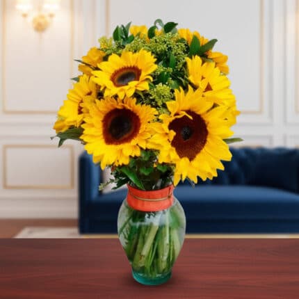 Arreglo de girasoles amarillos con follaje verde en un jarrón de cristal con cinta naranja, colocado sobre una mesa de madera frente a un sofá azul y una pared elegante