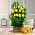 Arreglo floral vertical de rosas amarillas en base rústica de madera acompañado de caja de chocolates Ferrero Rocher sobre mesa de madera