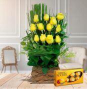 Arreglo floral vertical de rosas amarillas en base rústica de madera acompañado de caja de chocolates Ferrero Rocher sobre mesa de madera