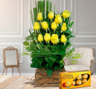 Arreglo floral vertical de rosas amarillas en base rústica de madera acompañado de caja de chocolates Ferrero Rocher sobre mesa de madera