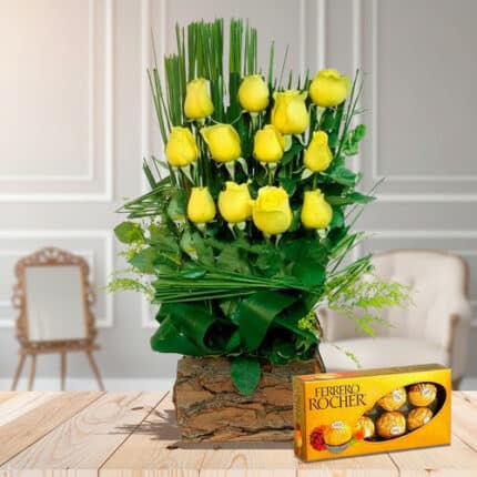 Arreglo floral vertical de rosas amarillas en base rústica de madera acompañado de caja de chocolates Ferrero Rocher sobre mesa de madera