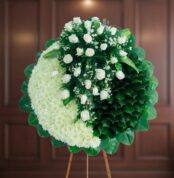 Arreglo floral redondo tipo corona con rosas blancas y hojas verdes sobre caballete de madera, fondo interior desenfocado