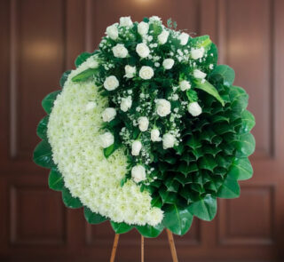 Arreglo floral redondo tipo corona con rosas blancas y hojas verdes sobre caballete de madera, fondo interior desenfocado