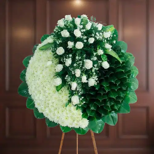 Corona floral redonda con rosas blancas y hojas verdes Arreglo floral redondo tipo corona con rosas blancas y hojas verdes sobre caballete de madera, fondo interior desenfocado