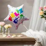 Globo metalizado en forma de estrella con la palabra feliz en colores vivos, colocado en un florero sobre una mesa con pétalos y rosas al fondo