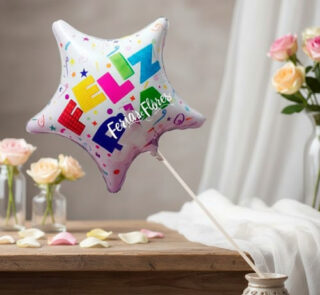 Globo metalizado en forma de estrella con la palabra feliz en colores vivos, colocado en un florero sobre una mesa con pétalos y rosas al fondo
