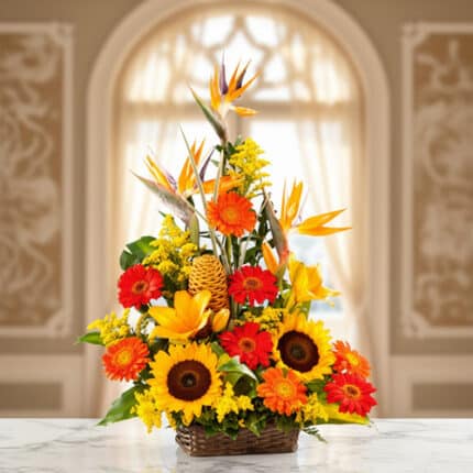 arreglo floral tropical con girasoles, gerberas naranjas, aves del paraíso y flores amarillas en canasta de mimbre sobre mesa de mármol