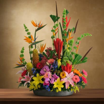 Centro de mesa con arreglo floral tropical de flores amarillas, rosas, rojas, naranjas y moradas en un jarrón bajo gris sobre una mesa de madera, con fondo marrón difuminado