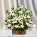 Arreglo de lirios y rosas blancas con follaje verde en base rústica de madera sobre mesa de mármol