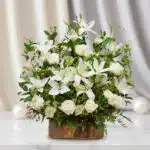 Arreglo de lirios y rosas blancas con follaje verde en base rústica de madera sobre mesa de mármol