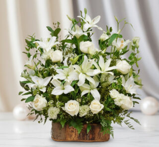 Arreglo de lirios y rosas blancas con follaje verde en base rústica de madera sobre mesa de mármol