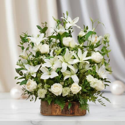 Arreglo de lirios y rosas blancas con follaje verde en base rústica de madera sobre mesa de mármol