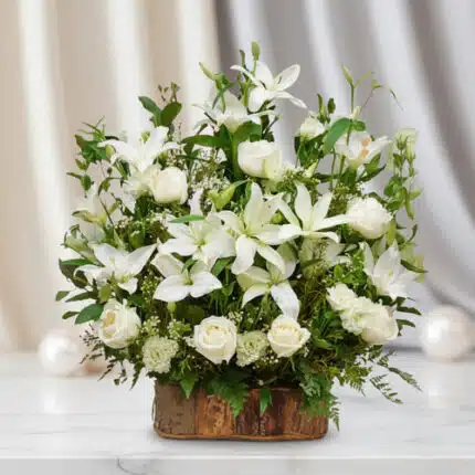Arreglo de lirios y rosas blancas con follaje verde en base rústica de madera sobre mesa de mármol