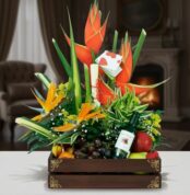 Arreglo floral tropical con heliconias y aves del paraíso acompañado de frutas variadas y una botella de vino en caja de madera decorativa sobre una mesa, con sala elegante y chimenea de fondo