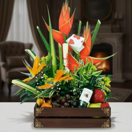 Arreglo floral tropical con heliconias y aves del paraíso acompañado de frutas variadas y una botella de vino en caja de madera decorativa sobre una mesa, con sala elegante y chimenea de fondo