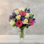 Arreglo floral con rosas blancas, gerberas rosas y flores moradas en un jarrón de cristal sobre superficie de mármol