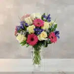 Arreglo floral con rosas blancas, gerberas rosas y flores moradas en un jarrón de cristal sobre superficie de mármol