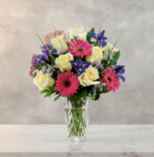 Arreglo floral con rosas blancas, gerberas rosas y flores moradas en un jarrón de cristal sobre superficie de mármol