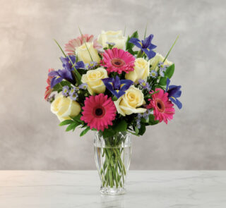 Arreglo floral con rosas blancas, gerberas rosas y flores moradas en un jarrón de cristal sobre superficie de mármol