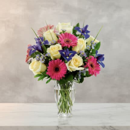 Arreglo floral con rosas blancas, gerberas rosas y flores moradas en un jarrón de cristal sobre superficie de mármol