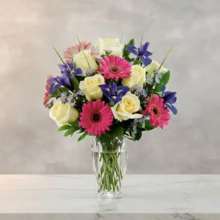 Arreglo floral con rosas blancas, gerberas rosas y flores moradas en un jarrón de cristal sobre superficie de mármol