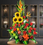 Arreglo floral grande con girasoles, rosas rojas, aves del paraíso y frutas como manzanas, uvas y naranja sobre mesa de madera con fondo desenfocado