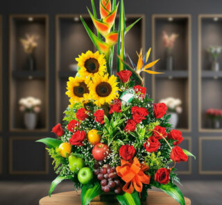 Arreglo floral grande con girasoles, rosas rojas, aves del paraíso y frutas como manzanas, uvas y naranja sobre mesa de madera con fondo desenfocado