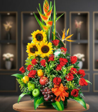 Arreglo floral grande con girasoles, rosas rojas, aves del paraíso y frutas como manzanas, uvas y naranja sobre mesa de madera con fondo desenfocado
