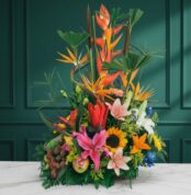 Arreglo floral tropical con heliconias, aves del paraíso, lirios, girasoles y frutas variadas sobre una mesa de mármol blanca frente a pared verde oscura